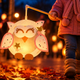 Kind hält eine leuchtende Eulen Laterne mit rosa Flügeln und goldenen Sternen am Laternenstab beim herbstlichen Laternenumzug am Abend