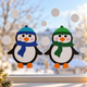 Zwei Pinguin Fensterbilder aus Tonkarton mit blauen und grünen Mützen und Schals hängen neben Schneeflocken am winterlichen Fenster.